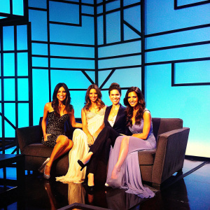 Giselle Blondet, Myrka Dellanos, Veronica Bastos, and Alix Aspe posing together in a chic studio setting. Wearing elegant dresses and suits. Cheerful expressions. Studio backdrop with geometric patterns and modern design.