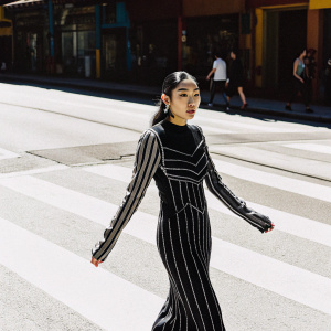 Serena, an Asian model, wearing a black and gray striped dress with intricate detailing. She stands confidently on a vibrant, sunlit street, surrounded by bustling city life.