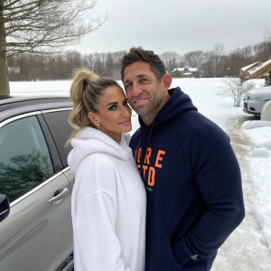Katie Price and Alex Reid at a snowy location near a parked car. Katie in a cozy white top and high ponytail. Alex in a navy hoodie with an orange logo. They share a warm, friendly pose.