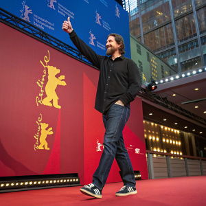 Christian Bale at the Berlinale Palast. Dressed in black shirt, jeans, and sneakers. The backdrop features the Berlin Film Festival logo in vibrant colors. He gestures with a thumbs-up sign.