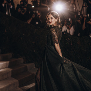 Emma Watson at the Met Gala, wearing a floor-length emerald green dress with intricate lace detailing. A sea of photographers and flashing lights fill the background.