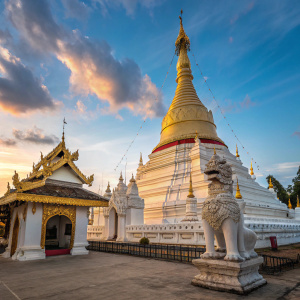In the heart of Mae Hong Son, Thailand, the grand temple Wat Phra That Doi Kong Mu rises with its majestic golden stupa. Intricate carvings decorate the structure, while a mythical guardian watches over the temple from the foreground. Above, the sky is painted with vibrant blue hues.