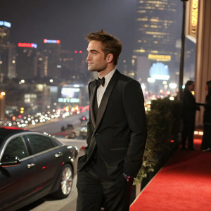 Robert Pattinson in a sleek black tuxedo, at the Golden Globe Awards, posing confidently against a bustling cityscape backdrop.
