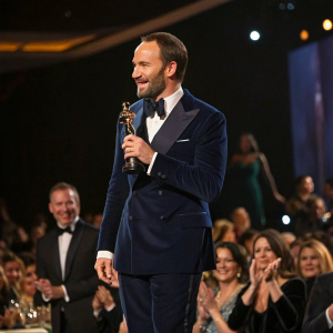 Tom Ford in a navy blue suit accepting an award at a lively gala. Crowd cheering in the background