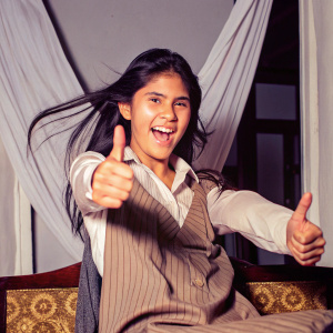 A vibrant image of a joyful Latina teen, giving a thumbs-up. She is dressed in a white shirt under a brown pinstriped blouse. Long dark hair flowing. The backdrop is a simple white curtain.