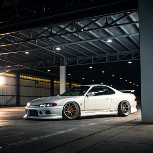 A sleek white Nissan Silvia S15 in an open garage, illuminated under dramatic lighting. The car's aerodynamic design and distinctive 18-inch alloy wheels are prominently featured. Shot from an angle that emphasizes the car's profile and modern lines.