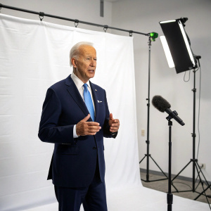 Joe Biden, in a dark suit and blue tie, addressing the press. White backdrop, studio lighting