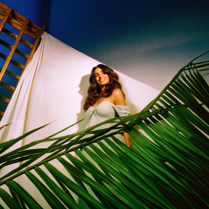 Monica Dogra, actress from Dhobi Ghat, posing in an elegant off-shoulder dress. She has long wavy dark brown hair and a warm smile. Dark studio backdrop