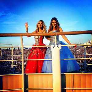 Lele Pons and Hannah Stocking attending the Radio Disney Music Awards. They are standing in front of a large crowd and flashing cameras. Lele, blonde hair, wearing a vibrant red dress with intricate gold detailing. Hannah, brunette, in a blue gown with sequins. Both are waving and smiling.