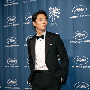 Wi Ha-jun, Korean actor, at Cannes Film Festival. Black tuxedo, white shirt, bow-tie. Blue backdrop with festival logos. Confident pose.