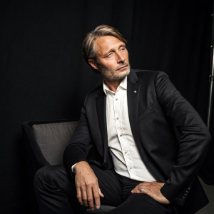 Close-up shot of Mads Mikkelsen. Wearing a sleek black blazer with white button-up shirt. Sitting in a minimalist studio with black backdrop. The lighting accentuates his sharp features and the texture of his attire.
