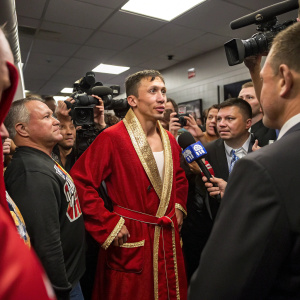 Gennady Golovkin standing in a packed locker room post-fight. Wearing a red robe with gold trim. Surrounded by a mix of excited supporters and formal-looking reporters with microphones.