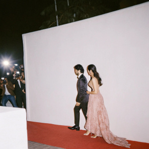 Kunal Nayyar and Neha Kapur attending a filmfare awards ceremony. Kunal in a sleek black tuxedo, Neha in a glamorous red gown. Red carpet event with flashing cameras