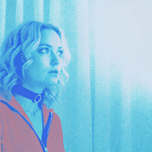 Close-up of Kate Hudson. Blonde wavy hair, red zipper jacket, black choker. Blurred white curtain background, spotlight