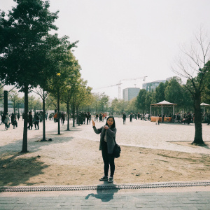 Nam Hong Joo, as portrayed by Bae Suzy, standing in a bustling city park. Wearing casual attire, she is waving towards the camera with a joyful expression.