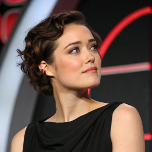 Close-up of Megan Boone, actress, with short, curly brown hair. Wearing a black dress. At a press conference. The background features a sleek, modern design with bold accents.