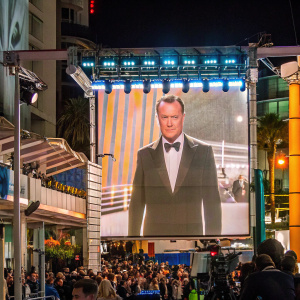 Gene Hackman, in sharp tuxedo, with stern look. Onstage at a film festival. Crowded, vibrant backdrop.