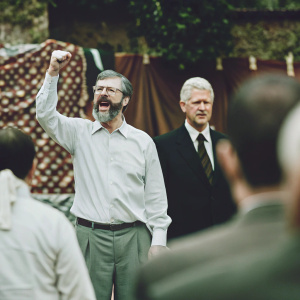 Gerry Adams, bearded and wearing glasses, making a passionate gesture. Behind him, Bill Clinton stands solemnly. Dark-suited figures gather around. The warm-hued tapestry serves as a backdrop.