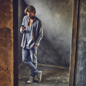 Jon Kortajarena, looking intense with tousled hair, in a casual outfit. He holds a smartphone casually. Standing solo in an empty, dimly lit room.