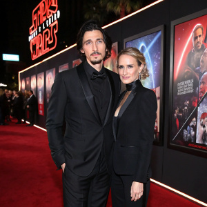 Adam Driver and Joanne Tucker at the Star Wars: The Last Jedi premiere. They are dressed formally in black suits. Surrounded by Star Wars movie posters with vibrant colors and stylized imagery.