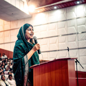 Malala Yousafzai, in a beautiful green saree, speaks at a TEDWomen conference. Bright stage lights, auditorium full of people.