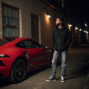 Vicky Kaushal, standing by a sleek red sports car. Poses in a dimly lit alleyway, wearing a casual outfit including a black hoodie and ripped jeans.