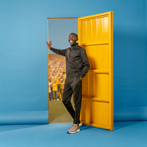 Ousmane Dembele, in a sleek black tracksuit, standing in a bustling stadium corridor. Crowd cheering in background.