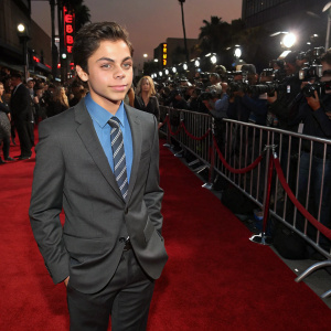 Jake T. Austin at a Hollywood premiere. Formal attire: gray suit, blue shirt, striped tie. Crowded red carpet with flashing cameras and paparazzi.