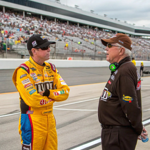 Kyle Busch and Joe Gibbs, both in racing gear, standing on the track at a NASCAR event. Crowd in the background.