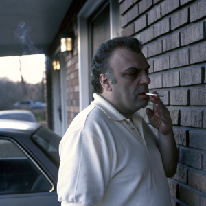 Close-up candid photo of Vincent Curatola, former 'The Sopranos' actor, smoking a cigarette. Wearing white polo shirt. Shot against a brick wall with natural lighting. Car partially visible in background.