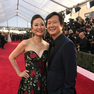 Tran Jeong and Ken Jeong at award show. Tran in a black floral gown, Ken in a dark suit. Crowded venue with photographers and press.