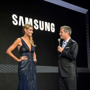 Heidi Klum and Tim O'Brian at the Samsung Electronics Unveiling Event. They stand in front of a sleek black backdrop with the Samsung logo. Both are dressed in formal attire.