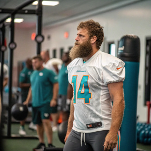Ryan Fitzpatrick, wearing Miami Dolphins jersey number 14, stands in a training session gym. Intense look on his face, preparing for a tough game