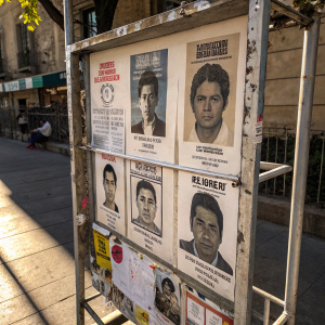 A poster with multiple black and white mugshots of individuals associated with the Arellano Felix drug cartel. The poster is mounted on a metal frame outdoors. It's covered with additional flyers and stickers. Bright sunlight casting shadows on the scene.