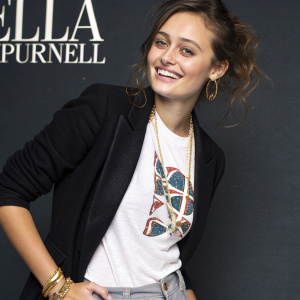 Ella Purnell, young actress with radiant smile, in casual chic outfit. White graphic tee, black blazer, gold accessories. Slightly messy hair. Dark backdrop with text elements. Studio portrait.
