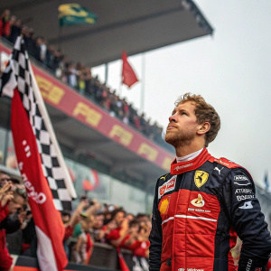 Sebastian Vettel, serious demeanor, standing in front of grand prix crowd. Wearing race suit with sponsor logos, amidst cheering fans.