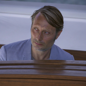 Caucasian actor Mads Mikkelsen at a film festival. Serious expression, stubble, short hair, wearing a blue blazer.