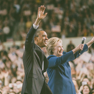 Barack Obama and Hillary Clinton, dressed in formal attire, addressing a cheering crowd at a rally. Obama in dark suit, Clinton in blue blazer, with raised arms. Crowd blurry in background.