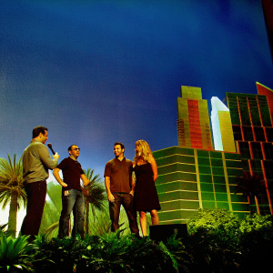 Tony Robbins, Gary Vaynerchuk, Sara Blakely, and Pitbull mingling at a vibrant tech conference. Futuristic cityscape backdrop