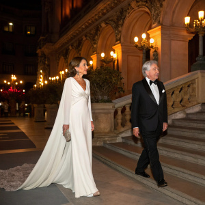 Isabel Preysler and Mario Vargas Llosa at an elegant evening gala. Isabel in a flowing white gown, Mario in a tailored tuxedo. Ornate venue with warm lighting.