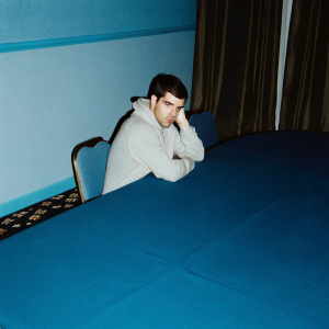 Simon Cowell, wearing a casual grey hoodie, seated at a large conference table. Multitude of business people in the background. Dimly lit room, conference event