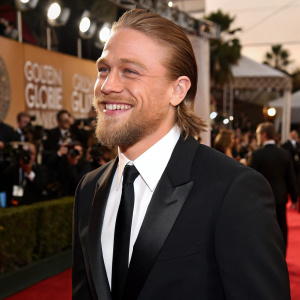 Charlie Hunnam, bearded and smiling, wearing a black tuxedo and tie. At the Golden Globe Awards on the red carpet. Paparazzi and celebrities in the background