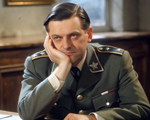 A German officer in uniform, with black hair parted in an undercut, sits at a desk, palm propped on his cheek.