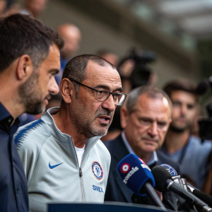 Maurizio Sarri addressing the media in a press conference. Flanked by assistants, behind them, blurred crowd of reporters.