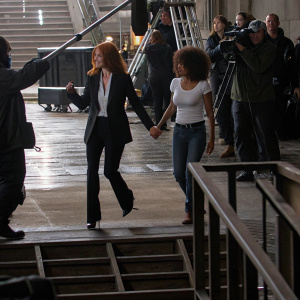Jessica Chastain and Halle Berry on a crowded movie set. Jessica in a stylish black suit, Halle in casual jeans and a white t-shirt. They're rehearsing a scene.