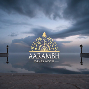 Aarambh Events Indore, logo design. Rich golden emblem centered. Decorative elements surround. Classic font choice. Indian cultural influence. Dark red backdrop