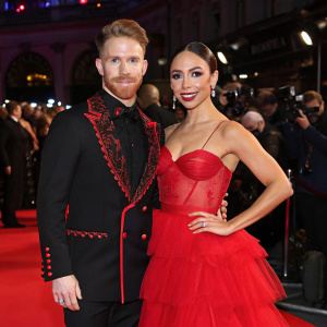 Neil Jones and Katya Jones at a gala event. He's in a black suit with red accents, she's in a red gown with layered tulle skirt. Crowds and cameras around them.