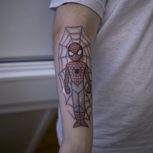 A detailed forearm tattoo featuring a web-slinging hero, inspired by Spider-Man, on a casual t-shirt wearing model.