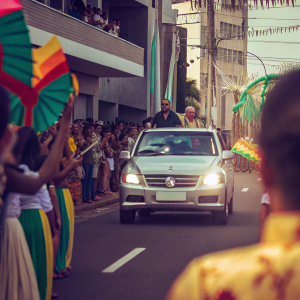 Pope Francis driving through a bustling street in a shiny car. Crowds of people in colorful attire lining the roads, waving flags. Festive atmosphere