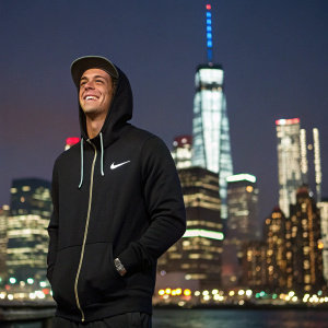 Ryan Sheckler, athlete, smiling for a magazine photo shoot. Wears black Nike hoodie. Behind him, bright city skyline